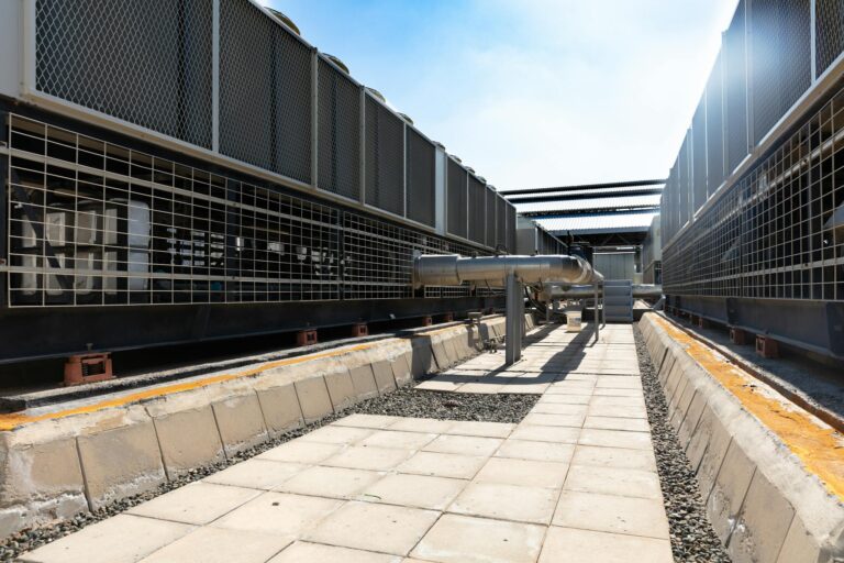 Detailed view of an industrial cooling system with metal pipes and sunlit concrete path.